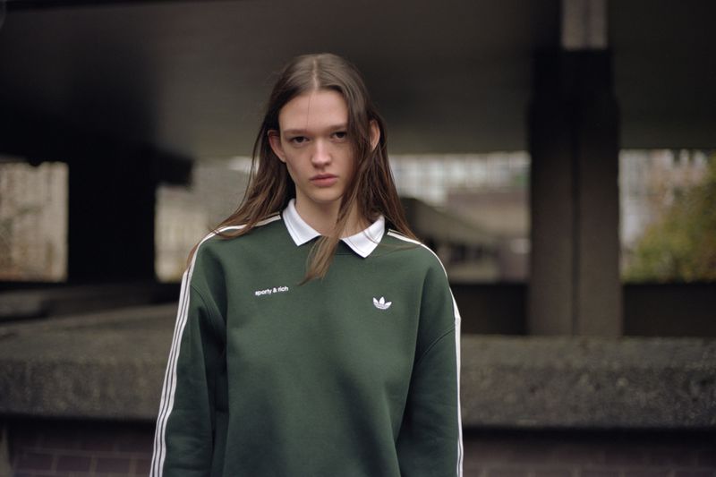 Adult female in sporty sweatshirt poses outdoors with neutral expression. Concrete and greenery are visible in background.