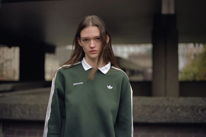 Adult female in sporty sweatshirt poses outdoors with neutral expression. Concrete and greenery are visible in background.