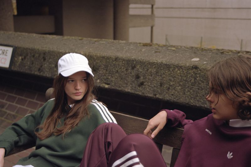 Two young adults sit on a concrete bench outside, wearing matching maroon and green Adidas tracksuits with a Brutalist architecture backdrop.