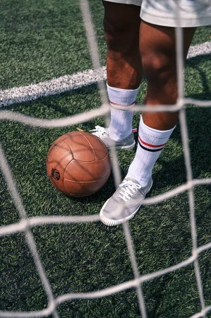 Person in athletic shoes and striped socks stands on a green field with a vintage brown leather soccer ball and a white net in front.
