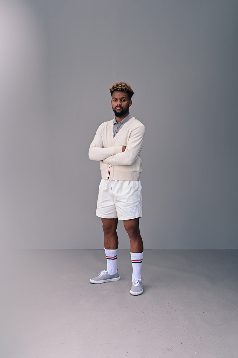 Full shot of a Black male in white shorts, socks, and sneakers with a light cardigan in front of a solid grey backdrop. 