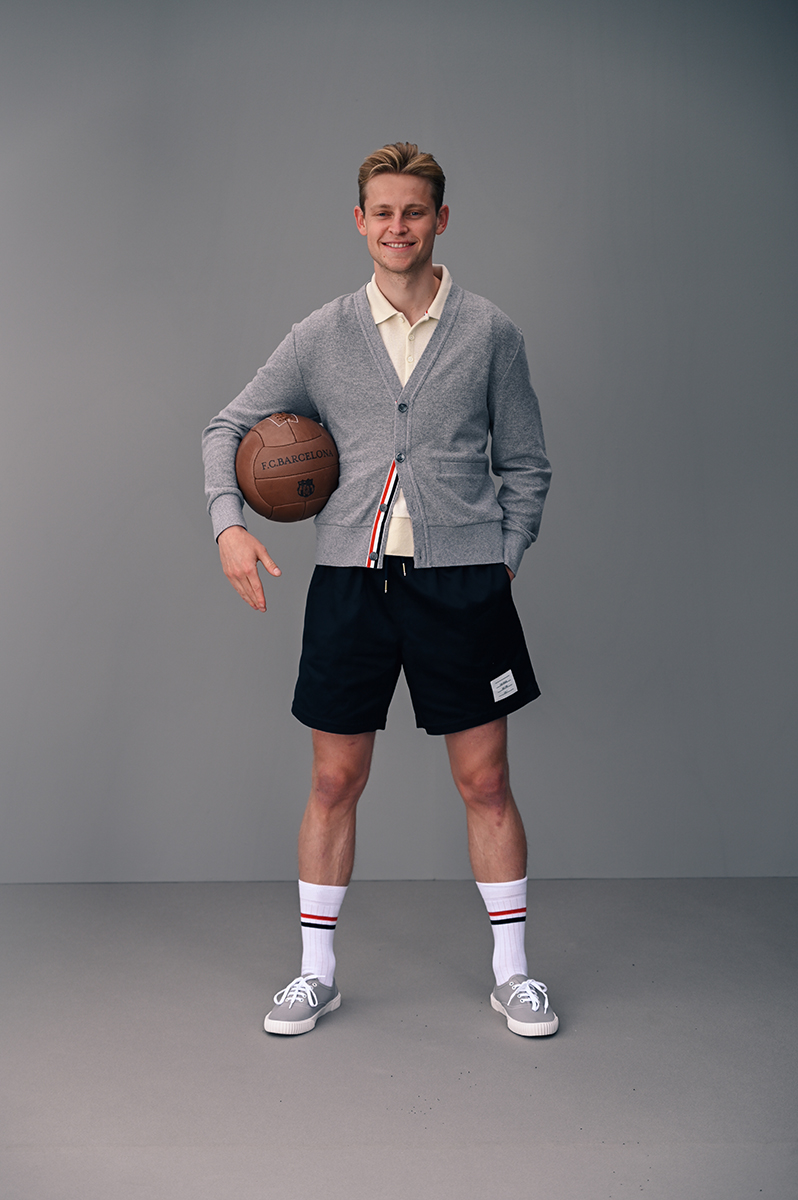 Full-shot of a smiling man in sportswear holding a soccer ball, set against a grey background in a studio setting. Fashion, sport.