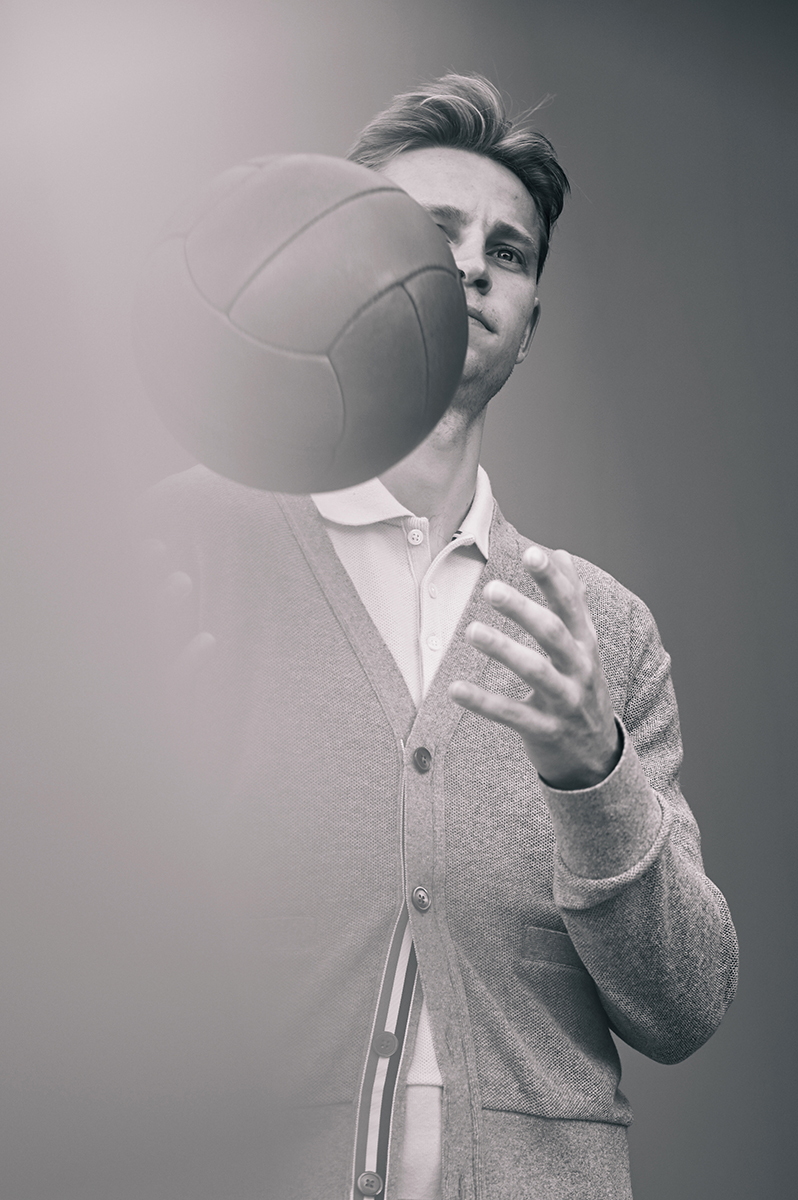 Monochrome portrait of a young man in a cardigan holding a volleyball, with a soft, blurred foreground and a gray backdrop.