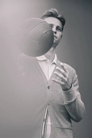 Monochrome portrait of a young man in a cardigan holding a volleyball, with a soft, blurred foreground and a gray backdrop.