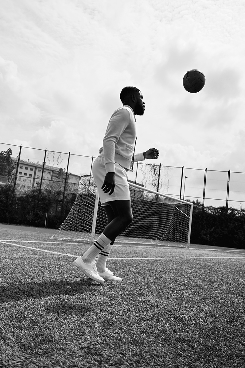 A Black male athlete in sportswear heading a soccer ball on a green field, overcast sky backdrop, promoting sports, fitness, and healthy lifestyle.