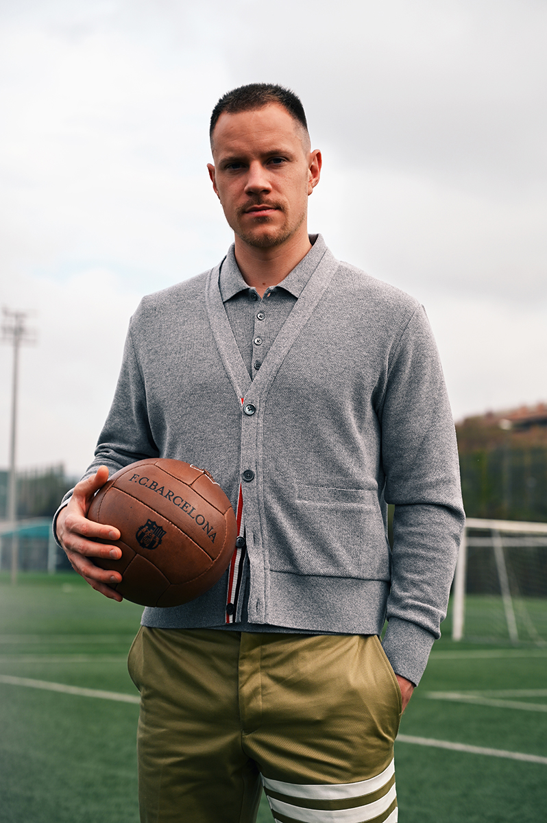 Man in gray cardigan and olive shorts holding a soccer ball, posing on a soccer field under an overcast sky; serious facial expression.