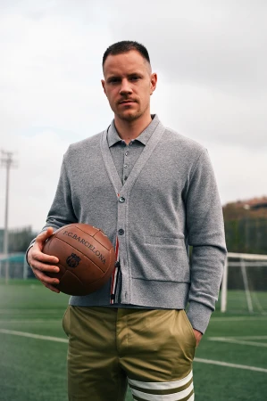 Man in gray cardigan and olive shorts holding a soccer ball, posing on a soccer field under an overcast sky; serious facial expression.