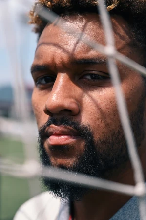 Close-up of a man with a beard and curly hair standing behind a white net with a blurred green and blue background on a sunny day.
