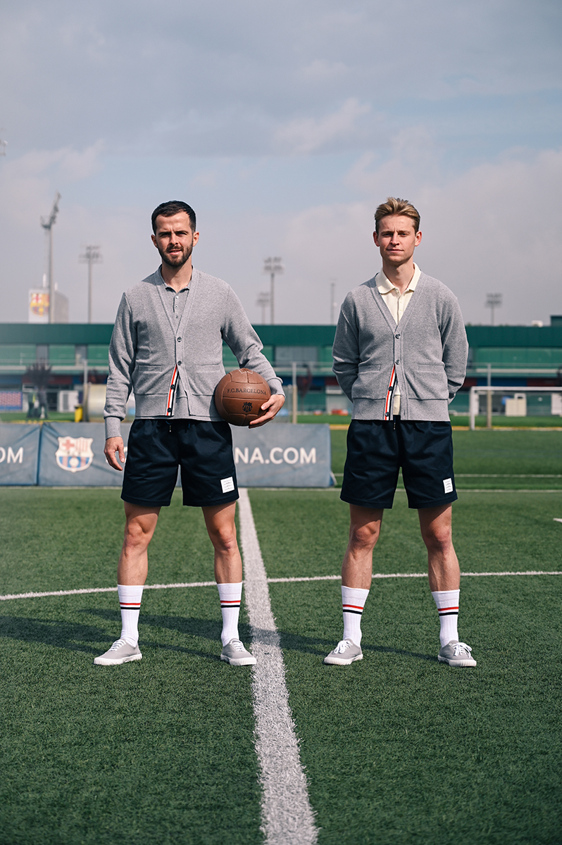 Two adult males in matching outfits stand on a soccer field, one holding a soccer ball, under a cloudy sky with stadium seating in background.