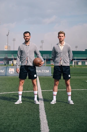 Two adult males in matching outfits stand on a soccer field, one holding a soccer ball, under a cloudy sky with stadium seating in background.