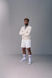 Man with folded arms wearing a cream cardigan, white shorts, and striped socks against a grey studio backdrop