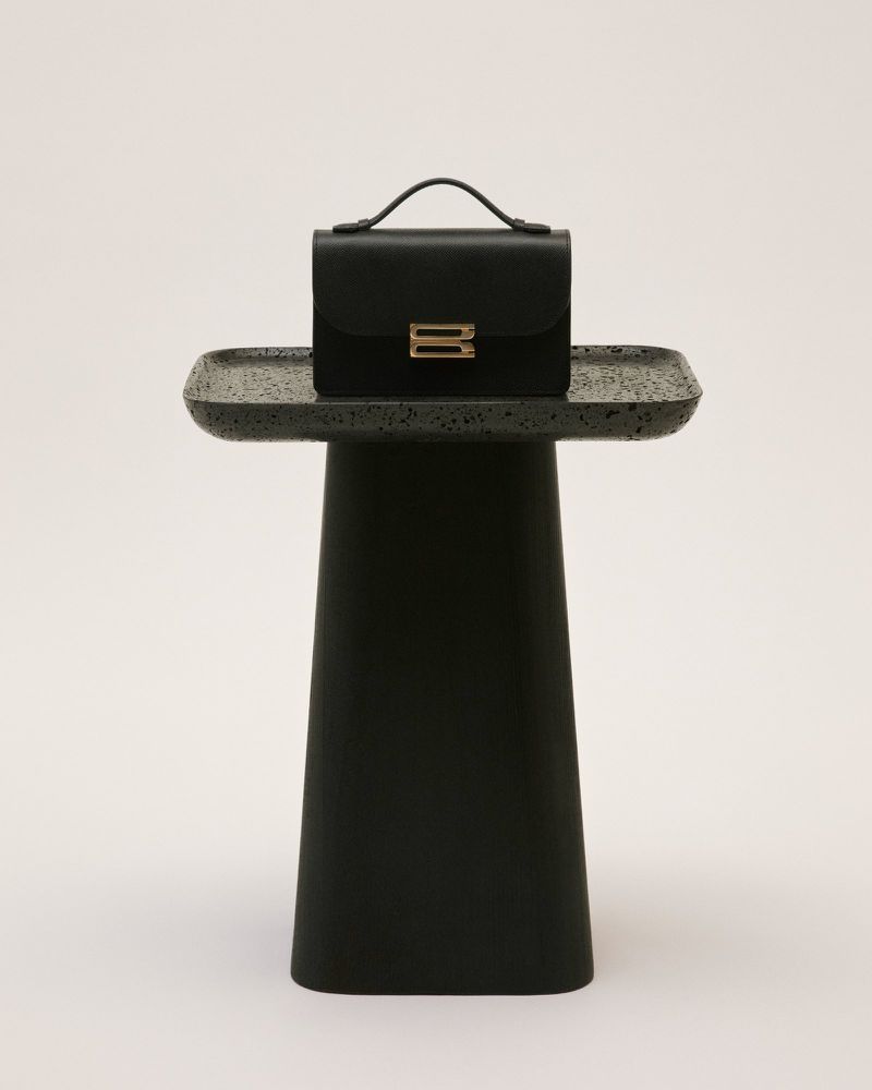 Black leather satchel handbag with gold hardware sitting on a black pedestal table against a clean, light background in a studio setting.