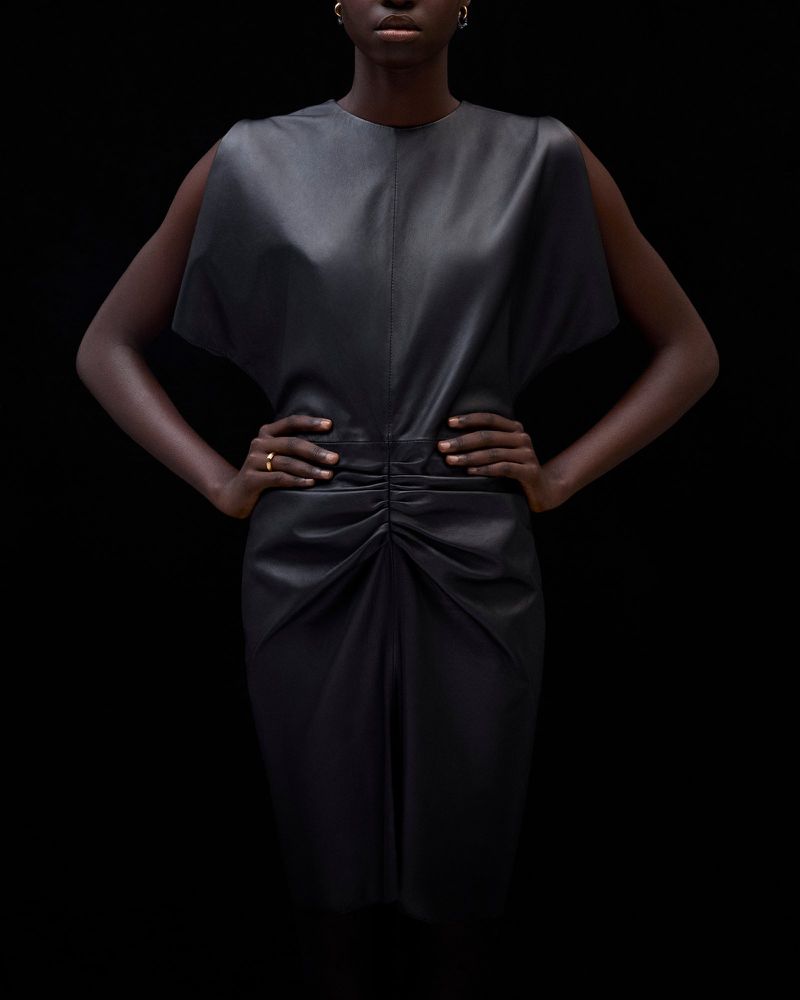 Fashionable woman in a black sleeveless dress with gathered waist detail, posed against a stark black backdrop in a studio setting.