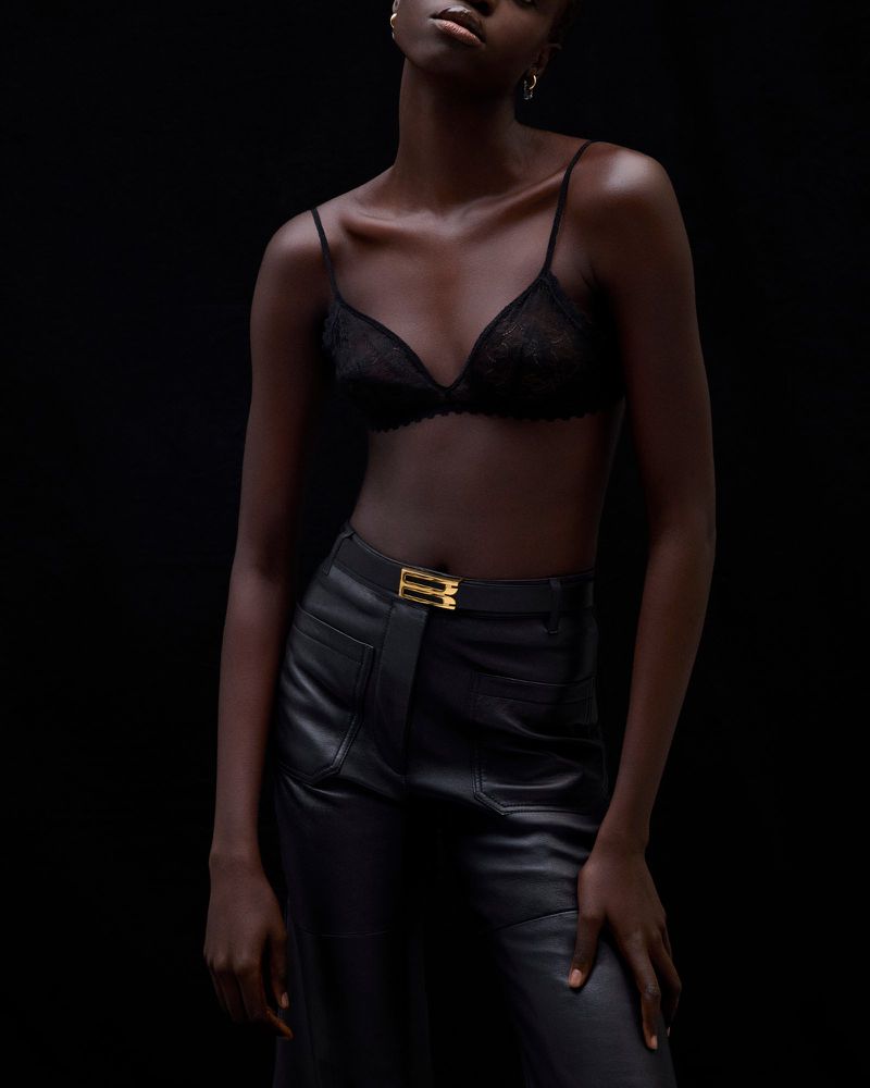Dark-skinned woman in a lace bra and leather pants poses against a black backdrop in a fashion studio shot.