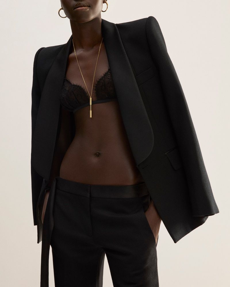 Fashionable dark-skinned woman in stylish black suit and bra, accessorized with gold necklace and earrings, against a neutral backdrop.