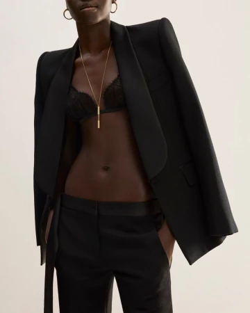 Fashionable dark-skinned woman in stylish black suit and bra, accessorized with gold necklace and earrings, against a neutral backdrop.