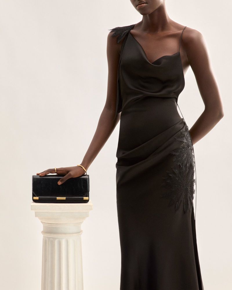 Elegant African-American woman in a black evening gown poses indoors with a black clutch on a white pillar, fashion portrait.