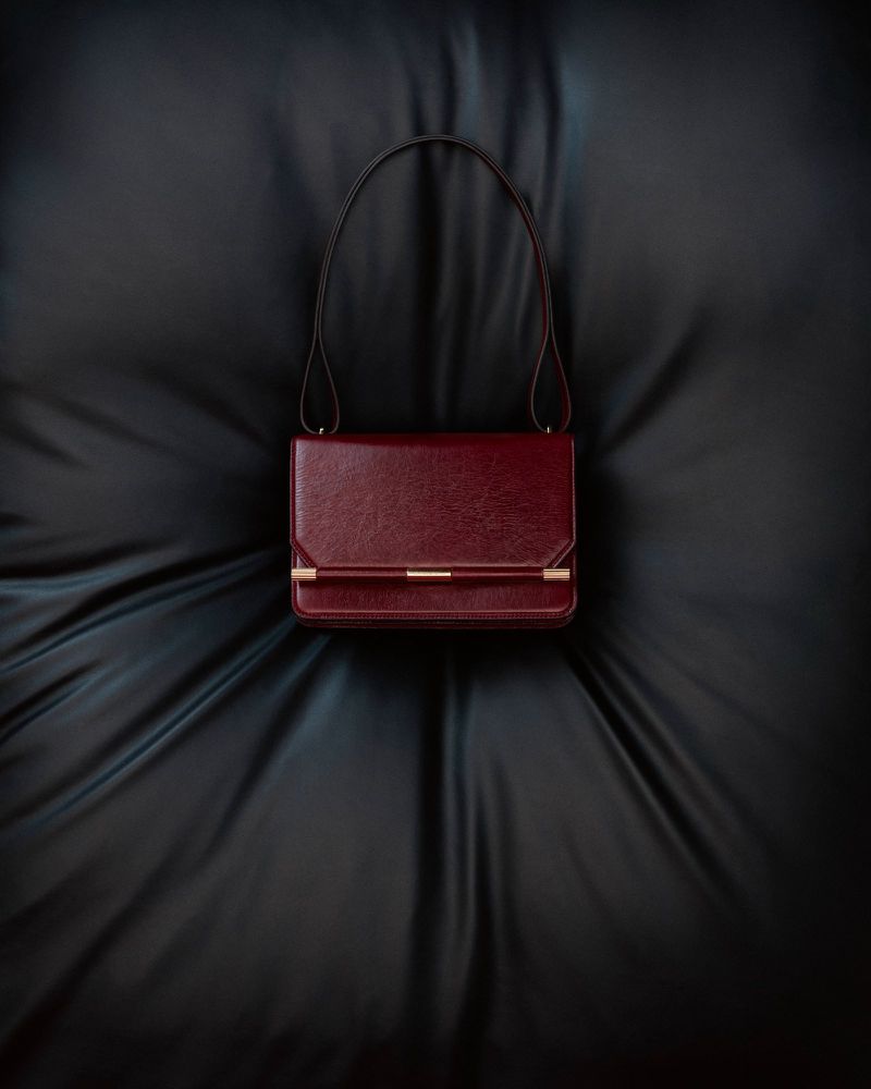 Burgundy leather shoulder bag with gold hardware sits on a draped black satin fabric, captured in a studio shot with centered composition.