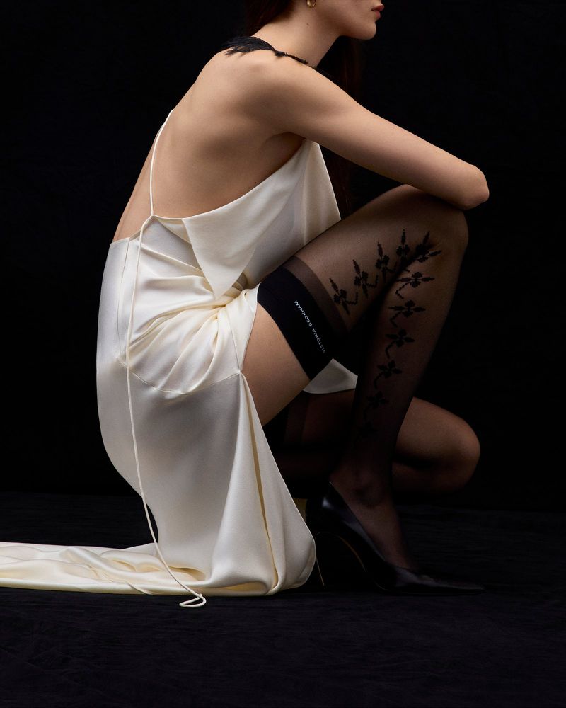 Woman in a cream-colored dress and stockings posing against a black backdrop, exhibiting a fashion-editorial aesthetic.