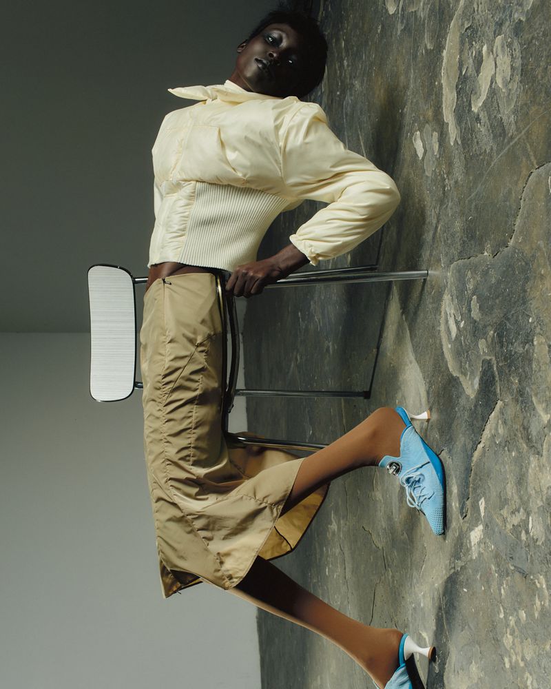 An African American woman leans against a chair, wearing a cream top, tan skirt, tights, and blue heeled shoes in a studio.