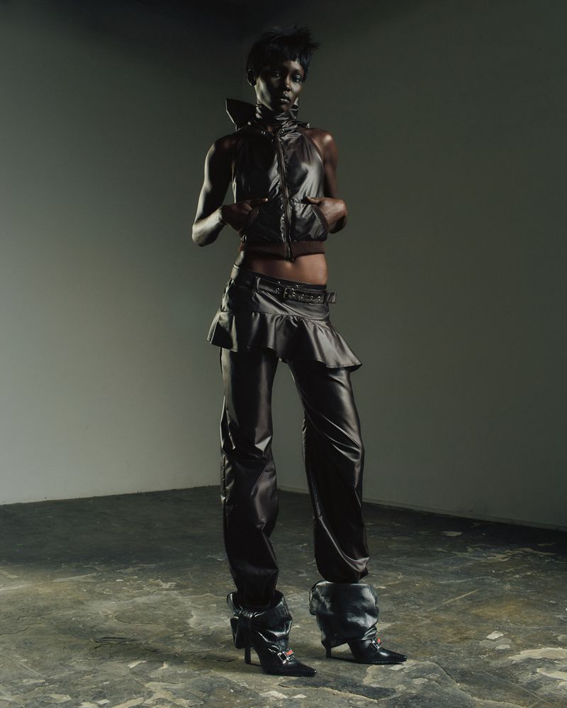 A Black fashion model in an edgy black leather outfit poses indoors with dramatic lighting, showcasing unique and modern fashion design.