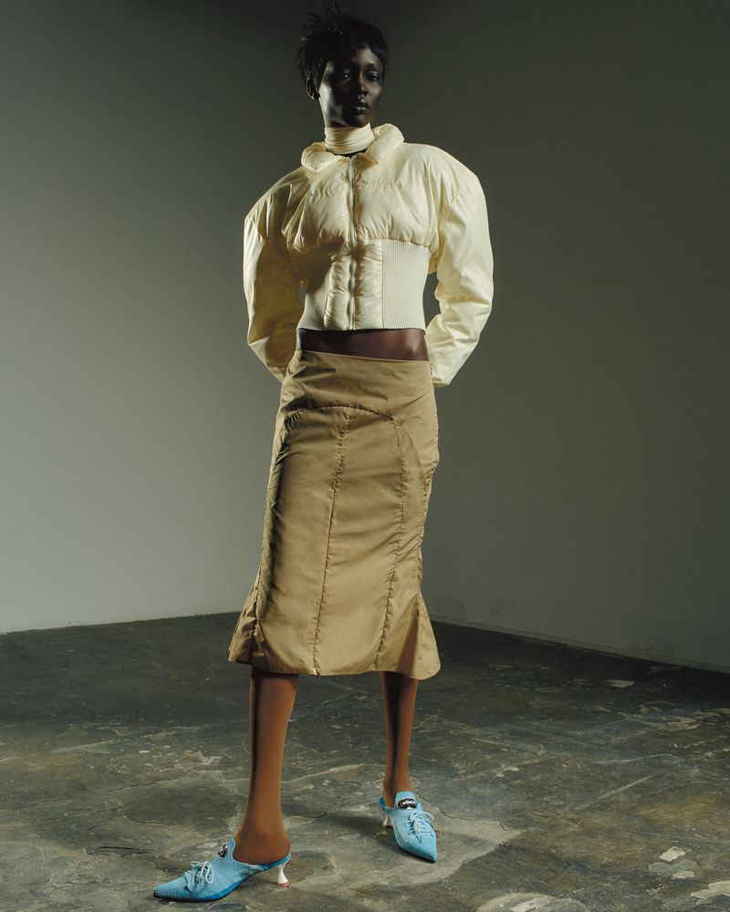Dark-skinned female fashion model in a short white puffer jacket, tan skirt, and blue heels in a studio with a gray backdrop.