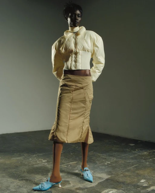 Dark-skinned female fashion model in a short white puffer jacket, tan skirt, and blue heels in a studio with a gray backdrop.