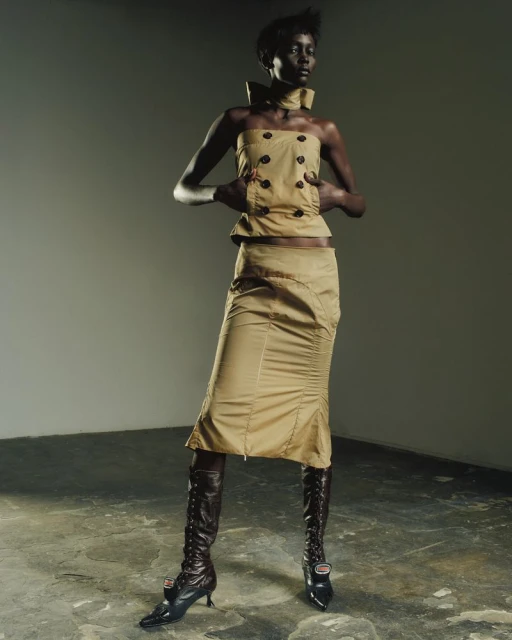 A dark-skinned woman poses in a tan strapless top and skirt with tall brown boots in a studio with a gray wall and concrete floor.