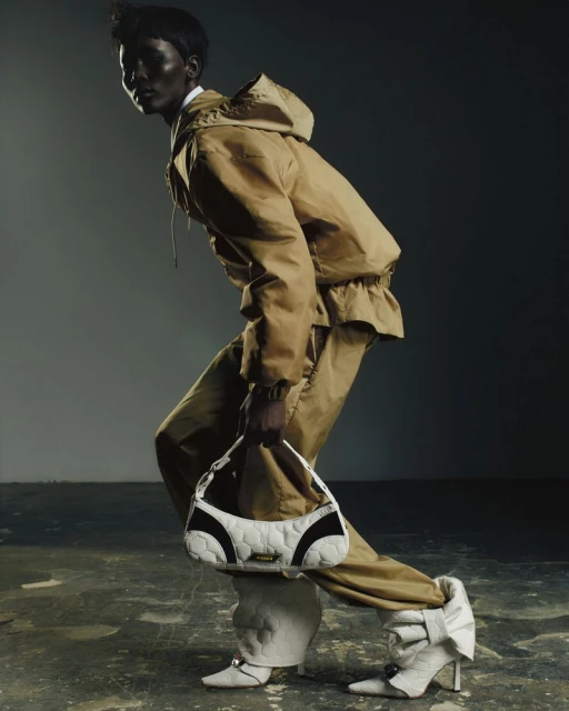 Fashion editorial shot of a Black model in a tan hooded suit and statement white heels, holding a white quilted handbag.