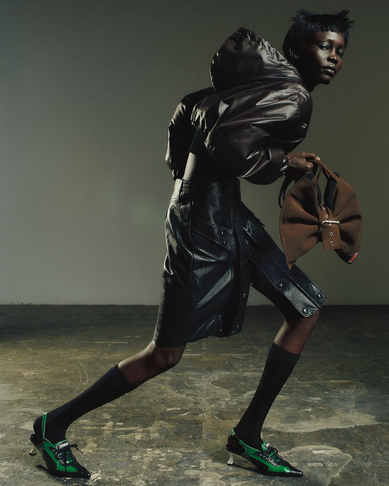 Fashion shot of a black female model wearing a black shiny jacket and shorts with green heeled shoes, holding a brown handbag.