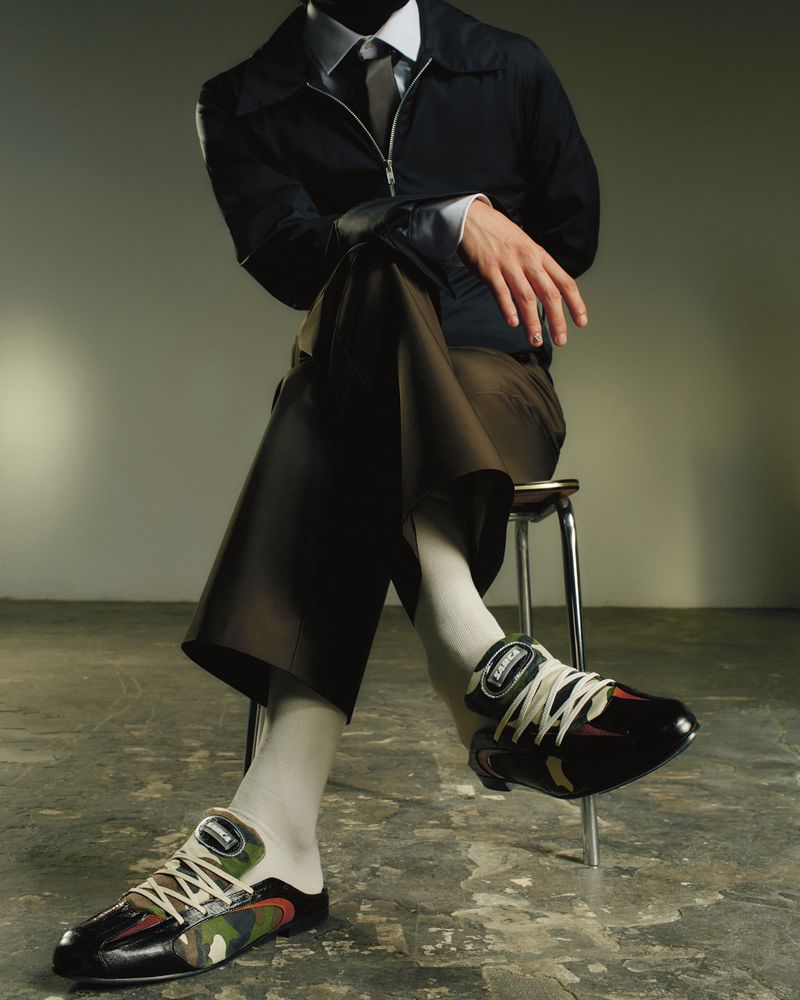 Man in dark suit and camouflage sneakers sits on stool against a grey studio backdrop, showcasing fashion and footwear.