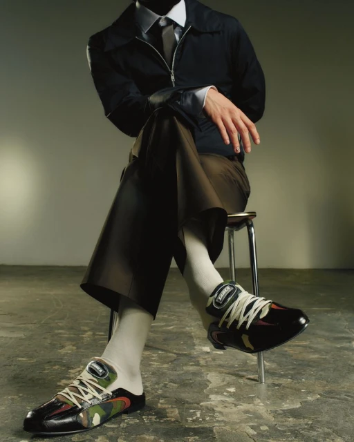 Man in dark suit and camouflage sneakers sits on stool against a grey studio backdrop, showcasing fashion and footwear.