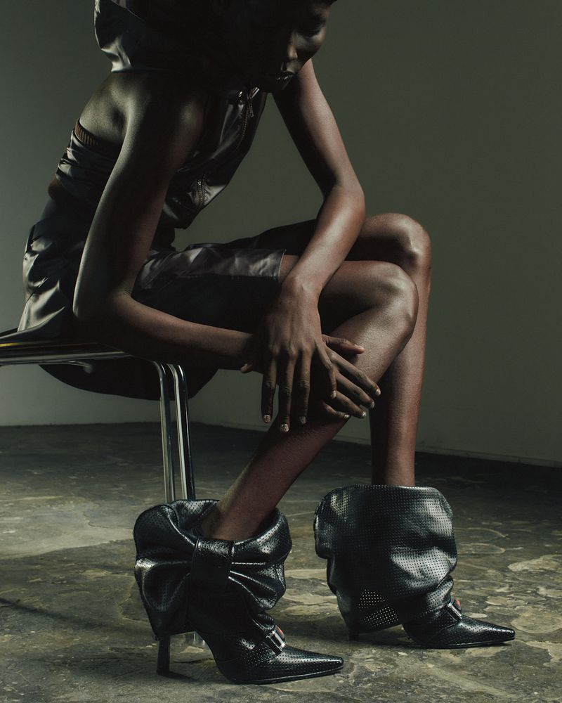 Dark-skinned model sits on metal chair wearing black textured mini dress and large black bow-detailed stiletto-heeled leather boots.