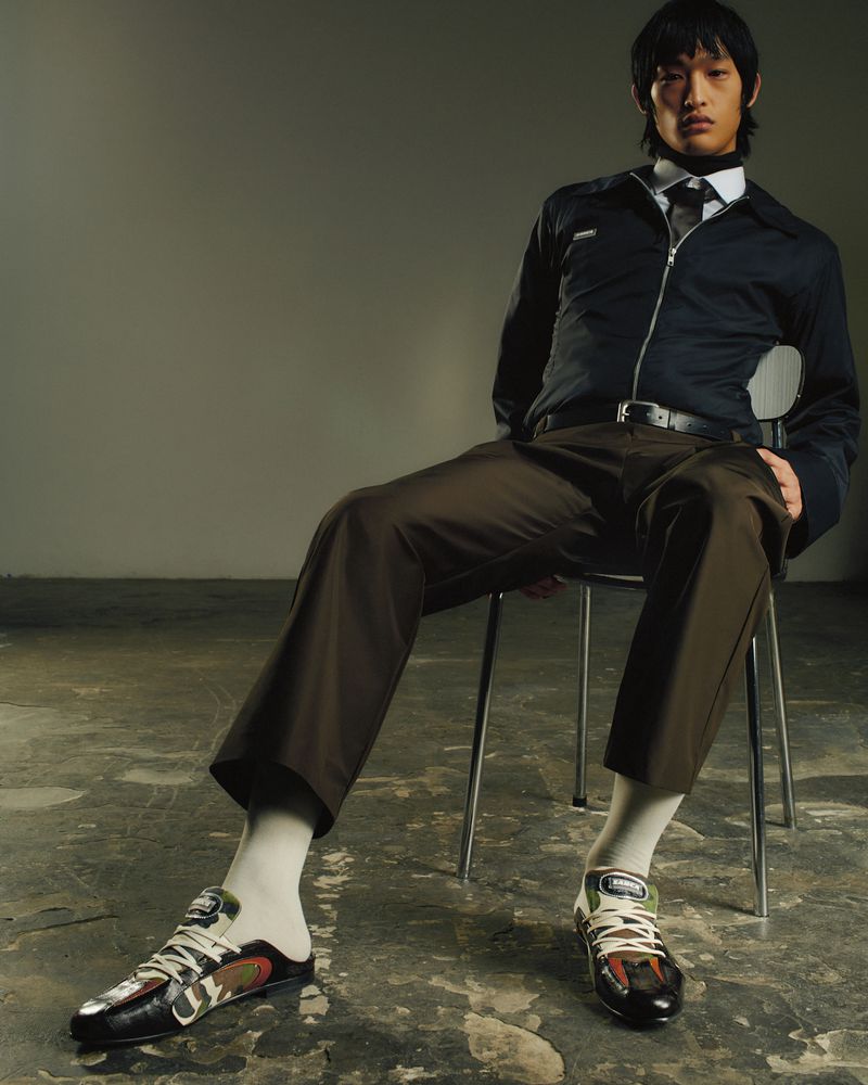 An Asian male fashion model sits on a metal chair wearing a navy jacket, dark pants, socks, and sneakers in a photo studio.