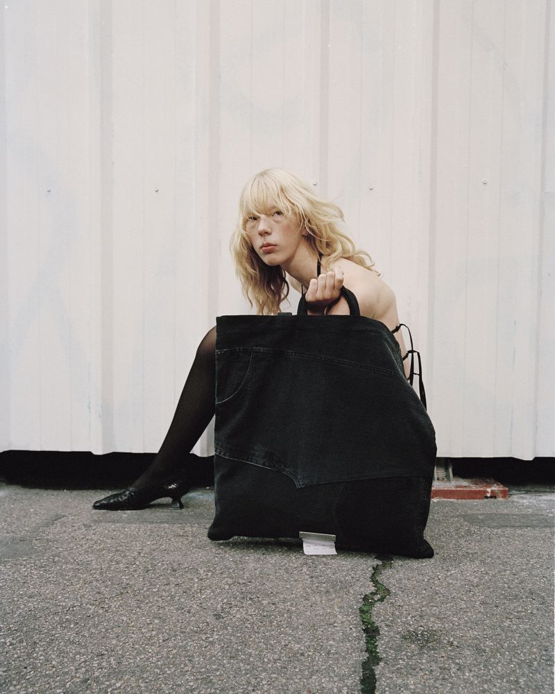 Model with blonde hair posing on pavement holding a large black denim tote bag against a pale vertical panel background