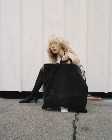 Model with blonde hair posing on pavement holding a large black denim tote bag against a pale vertical panel background