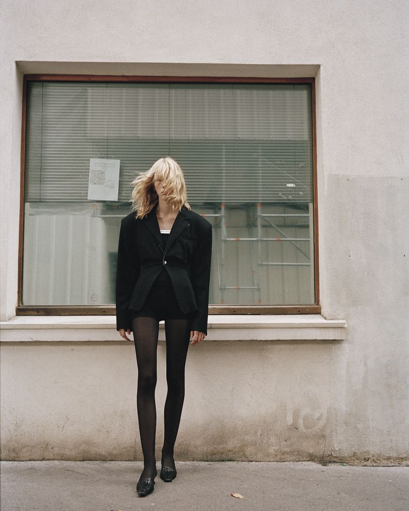 A blonde woman stands outside a building with a window wearing a black blazer, shorts, tights, and shoes in a fashion editorial.