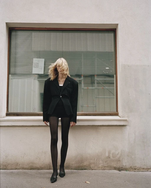 A blonde woman stands outside a building with a window wearing a black blazer, shorts, tights, and shoes in a fashion editorial.