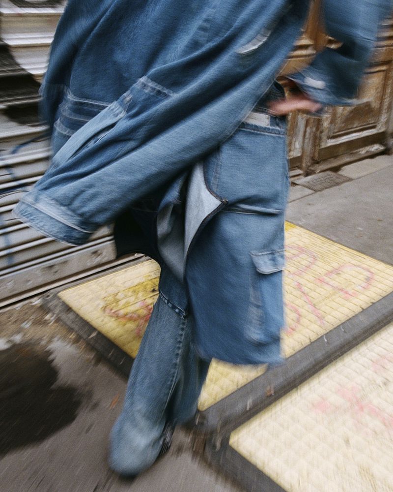 A person walking on a street in denim clothing, with a jacket draped over their shoulder, motion blur effect.