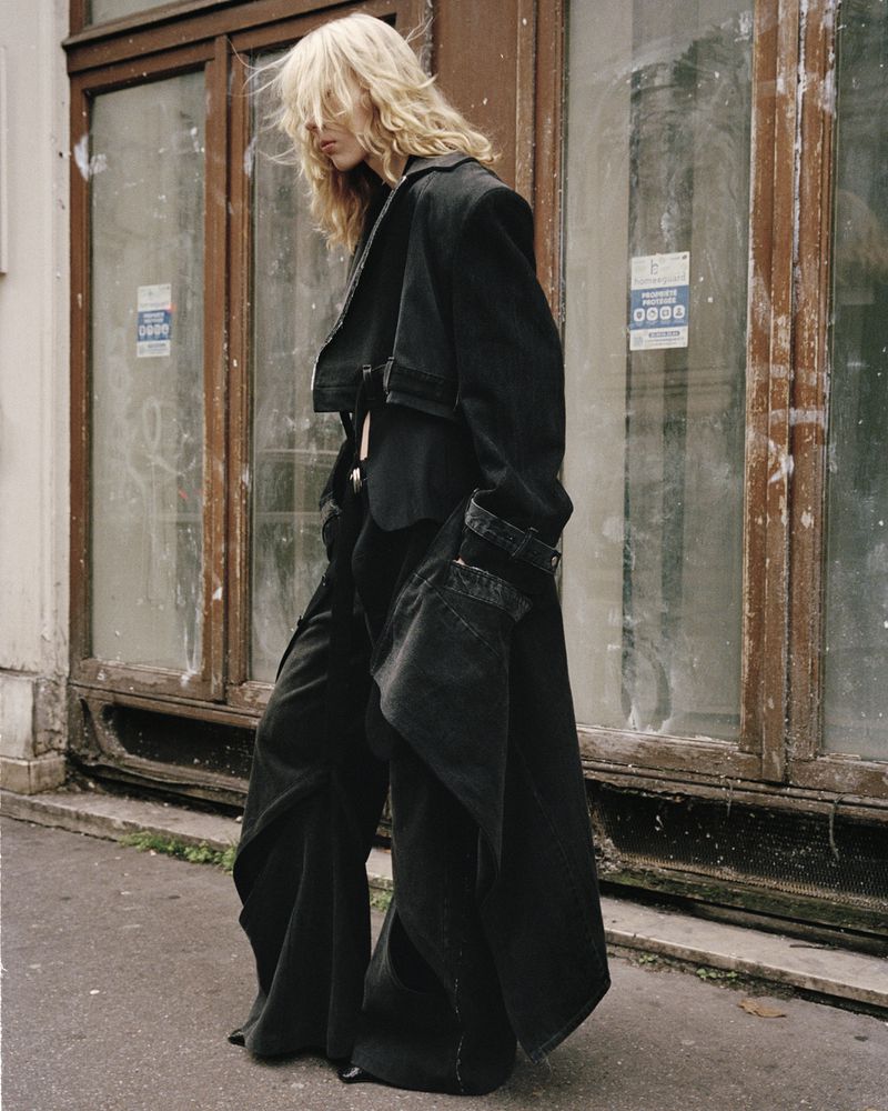 A woman stands in a wide-legged pantsuit and long coat. She's outdoors in front of an old building with weathered windows, creating an urban fashion shot.