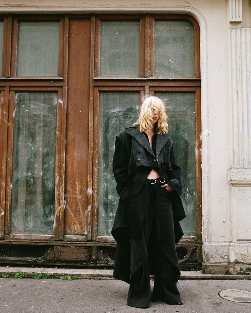 A young adult woman with blonde hair in a black jacket and pants stands in front of weathered brown doors on a city street.