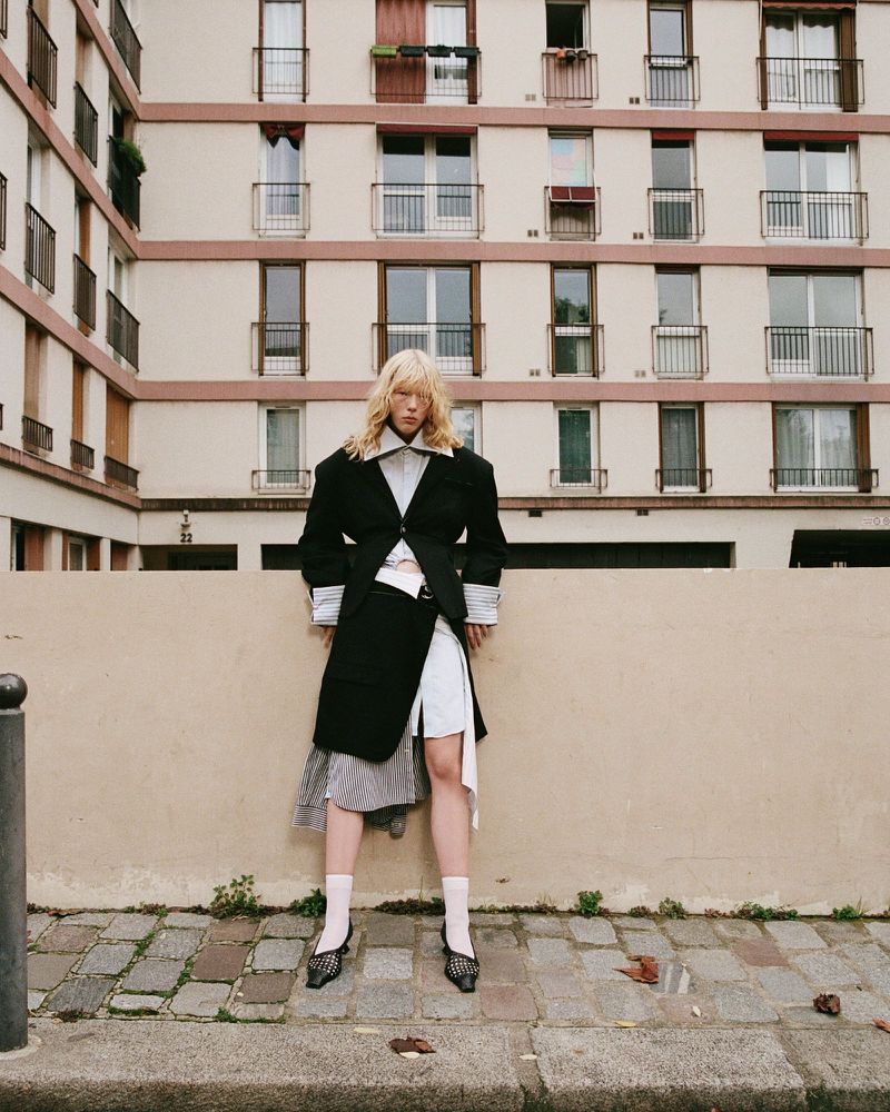 Fashionable woman in black blazer poses against tan wall in front of apartment building on paved street wearing white socks and heels.