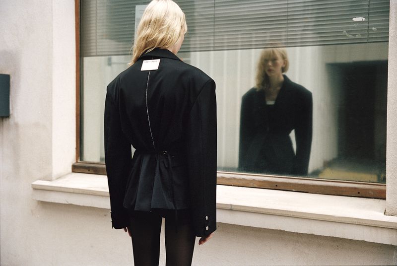 Blonde model seen from behind wearing a black oversized blazer jacket, reflected in a large street-facing window.