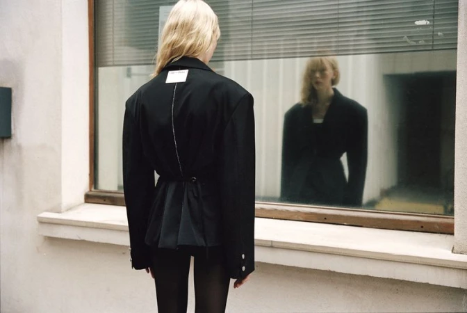Blonde model seen from behind wearing a black oversized blazer jacket, reflected in a large street-facing window.