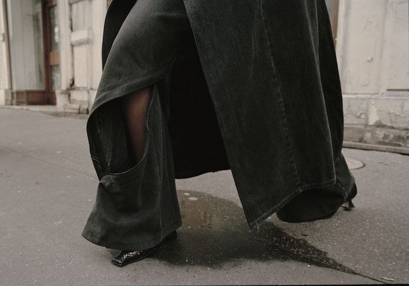 Person wearing grey, wide-leg jeans and black heels walking on a city street with blurred building and sidewalk in the background.