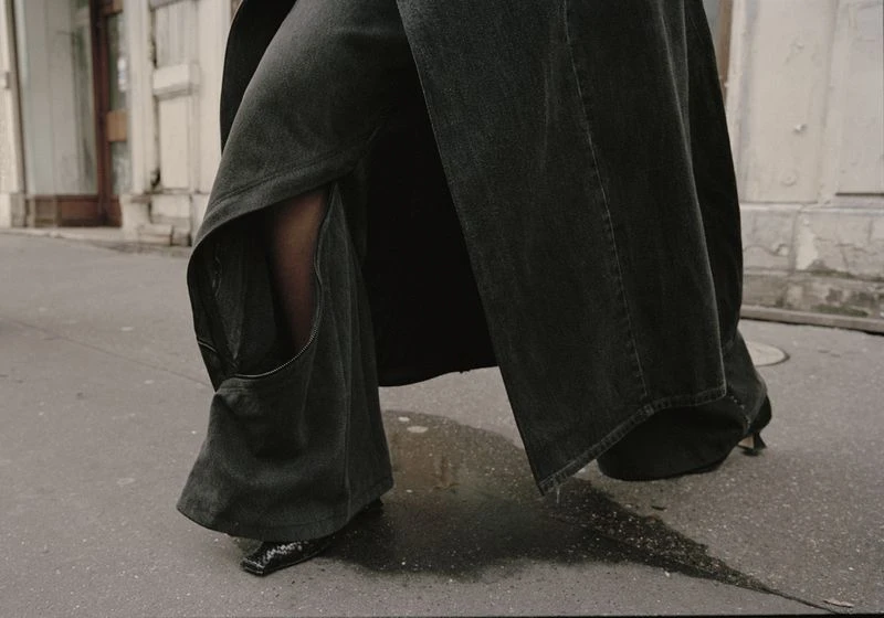 Person wearing grey, wide-leg jeans and black heels walking on a city street with blurred building and sidewalk in the background.