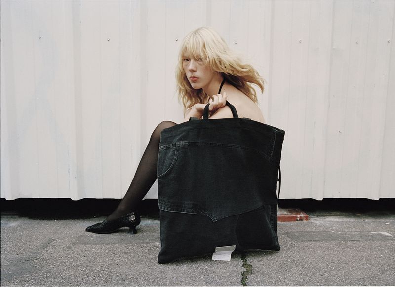 Blonde woman sits beside a large black denim tote bag against a white corrugated wall; she wears black tights and heeled shoes.