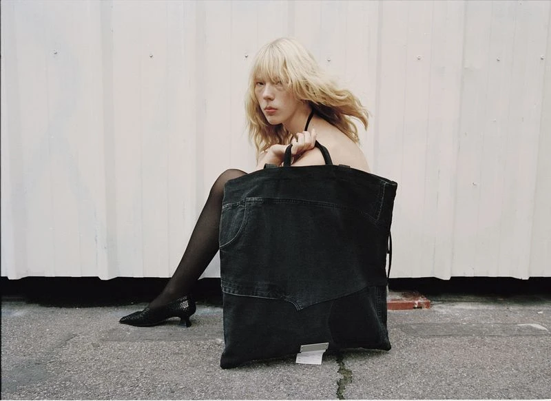 Blonde woman sits beside a large black denim tote bag against a white corrugated wall; she wears black tights and heeled shoes.