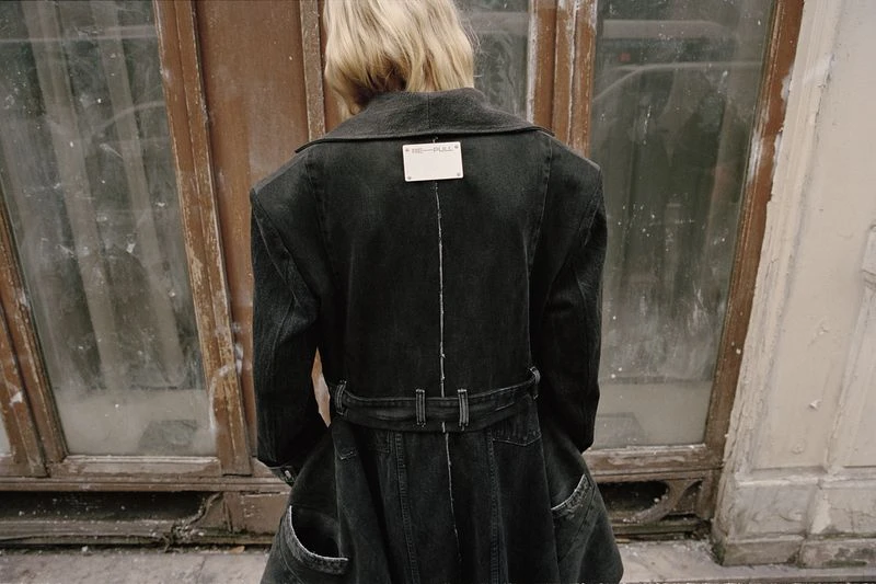 Back view of a blond person in a dark denim jacket dress standing in front of an old weathered door with window panes.