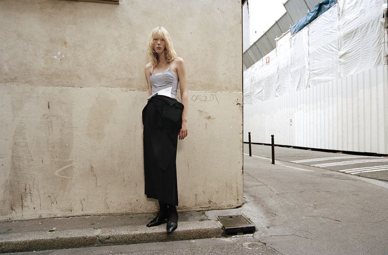 Fashion shot of a blond female model in a grey top and black skirt, posing against a textured beige wall in an urban environment.